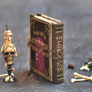 May include: A miniature, handcrafted book with a dark brown cover, secured by a chain. A skull-shaped candle holder, a small green bottle, a cork, and two small bones are also present, creating a gothic theme.