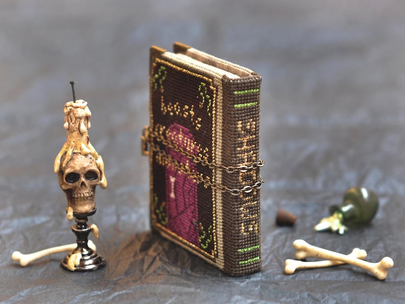 May include: A miniature, handcrafted book with a dark brown cover, secured by a chain. A skull-shaped candle holder, a small green bottle, a cork, and two small bones are also present, creating a gothic theme.