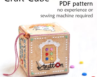 May include: A beige fabric cube with a cross-stitch design of a button on one side. The cube has a pink string attached to it. The text "Craft Cube PDF pattern no experience or sewing machine required" is above the cube. The text "button" is stitched on the cube.