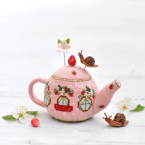 May include: A pink crocheted teapot pincushion with a strawberry on top and two snails on the side. The teapot has three windows with red and blue accents. The pincushion is sitting on a white wooden surface.