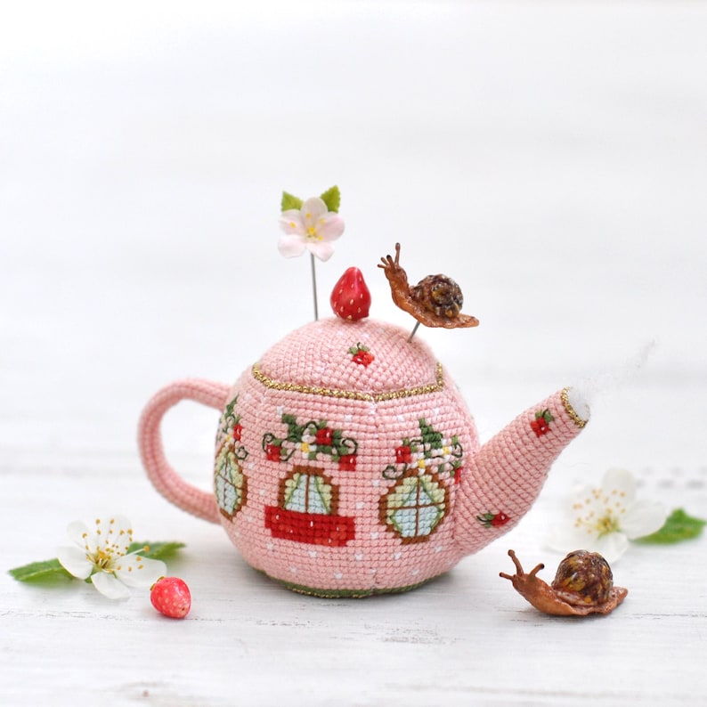 May include: A pink crocheted teapot pincushion with a strawberry on top and two snails on the side. The teapot has three windows with red and blue accents. The pincushion is sitting on a white wooden surface.