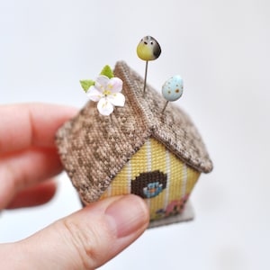 May include: A small yellow and brown birdhouse pincushion with a white flower and blue egg accents. The pincushion is made of fabric and has a brown roof and yellow walls.