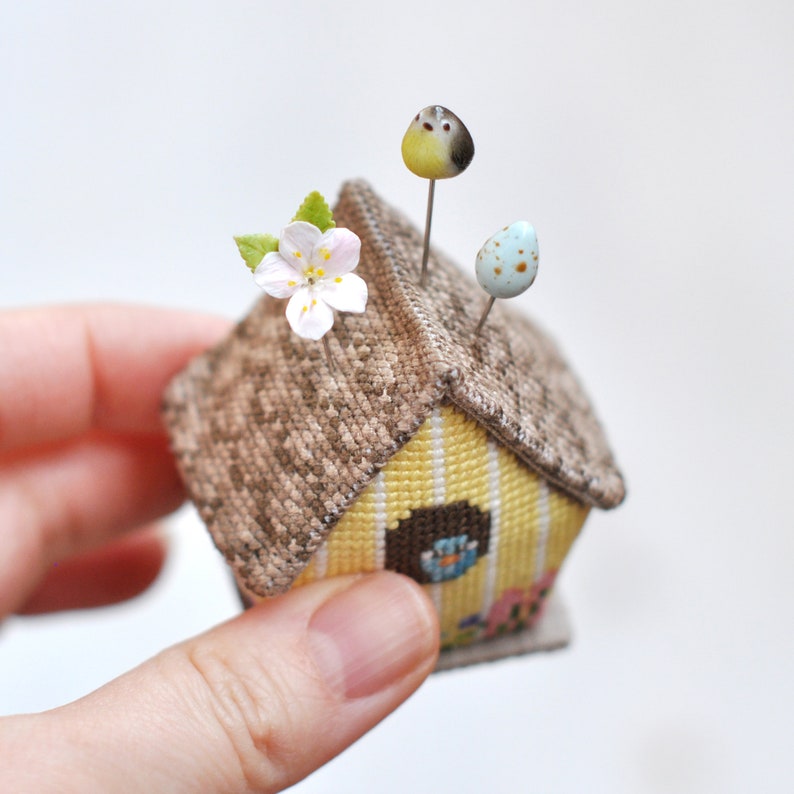 May include: A small yellow and brown birdhouse pincushion with a white flower and blue egg accents. The pincushion is made of fabric and has a brown roof and yellow walls.