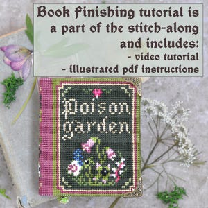 Op de afbeelding: Een klein, met de hand gestikt boek getiteld "Poison Garden" met een roze en groene omslag. Het boek is versierd met een kruissteekontwerp van bloemen en een zwarte kat.