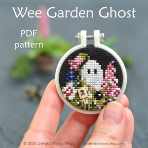 Könnte beinhalten: Ein Kreuzstich-Stickrahmen mit einem weißen Geist, schwarzen Augen und einem Schild mit Totenkopf und gekreuzten Knochen. Das Design umfasst rosa, blaue und grüne florale Elemente auf schwarzem Hintergrund. Der Text oben lautet "Wee Garden Ghost PDF pattern."