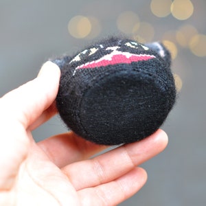 May include: Black knitted plush toy with a red and white embroidered design. The toy is shaped like a small pot or container.