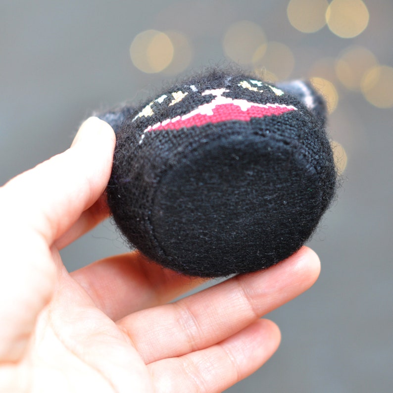 May include: Black knitted plush toy with a red and white embroidered design. The toy is shaped like a small pot or container.