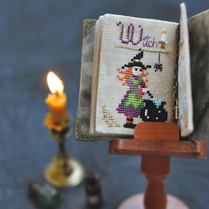May include: A miniature cross-stitch book with a witch theme. The cover features the word "Witch" and a stitched image of a witch stirring a cauldron. The book is displayed on a small wooden stand, with a lit candle in the background.