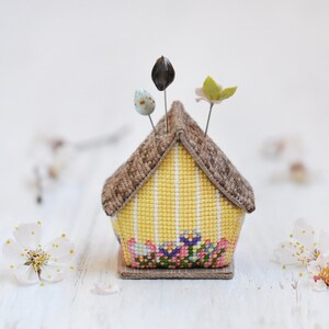 May include: A yellow and brown house-shaped pincushion with a floral design and three pins. The pincushion is sitting on a white surface.
