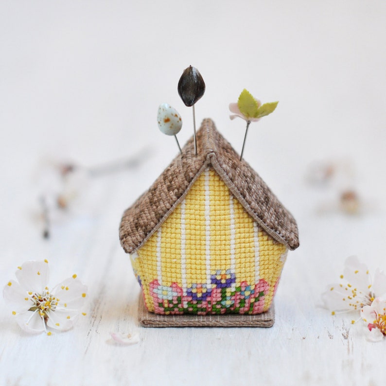 May include: A yellow and brown house-shaped pincushion with a floral design and three pins. The pincushion is sitting on a white surface.