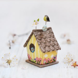 May include: A yellow birdhouse pincushion with a brown roof, a small bird on top, and a flower on the side. The pincushion is decorated with a floral pattern and has a small base.