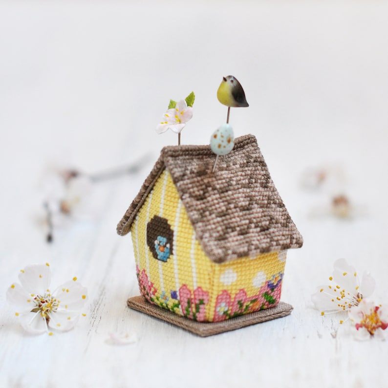 May include: A yellow birdhouse pincushion with a brown roof, a small bird on top, and a flower on the side. The pincushion is decorated with a floral pattern and has a small base.
