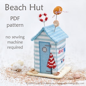 Op de afbeelding: Een blauw-wit gestreept strandhuisje-naaldenkussen met een rood-wit gestreepte parasol, een rode reddingsboei, een schelp en een gele bloem erop. Het naaldenkussen staat op een basis van schelpen. De tekst "Beach Hut" staat bovenaan de afbeelding en de tekst "PDF pattern" eronder. De tekst "no sewing machine required" staat eronder.