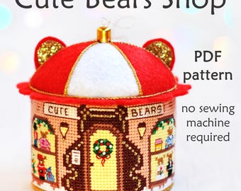 Può includere: Un ornamento natalizio in feltro rosso e bianco a forma di un negozio di orsacchiotti. L'ornamento ha un cartello che dice "Cute Bears Shop" e presenta una porta con una ghirlanda. L'ornamento è decorato con glitter dorati e ha un nastro dorato.