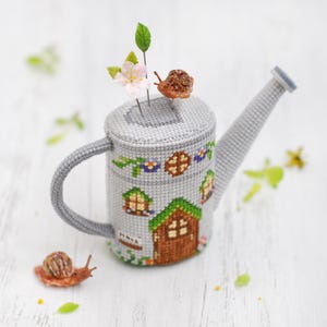 K&ouml;nnte beinhalten: Eine graue Keramik-Gie&szlig;kanne mit einem Kreuzstich-Design eines Hauses und Blumen. Die Gie&szlig;kanne hat einen braunen Griff und eine kleine Schnecke oben drauf.
