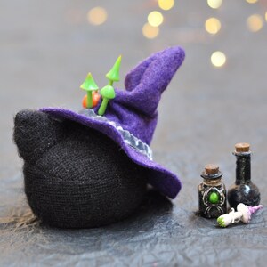 May include: A black knitted witch with a purple hat and green mushroom accents. The witch is holding two small glass bottles with green and black liquid.