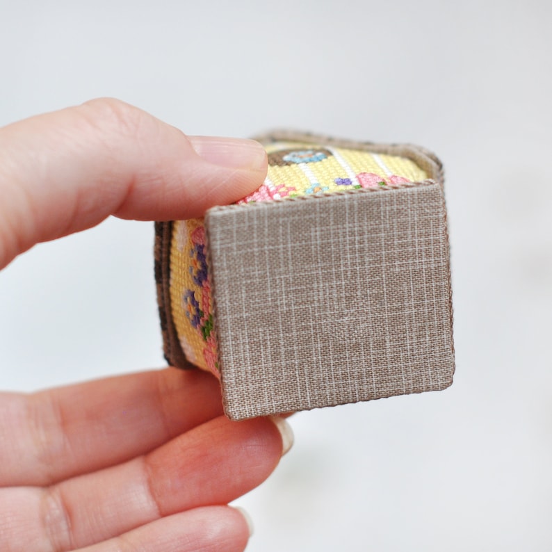 May include: A small, square-shaped box with a floral pattern and a brown fabric lining. The box is made of fabric and has a yellow, pink, and white floral design.