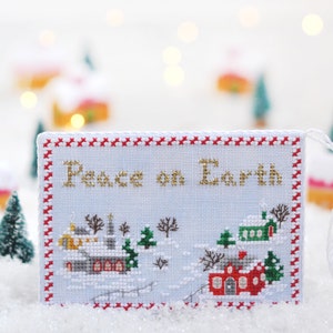 May include: A white cross-stitch ornament with a red and white border. The ornament features a winter village scene with the text "Peace on Earth" in gold thread.