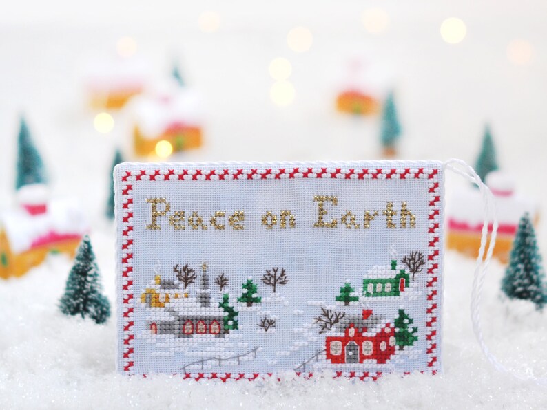 May include: A white cross-stitch ornament with a red and white border. The ornament features a winter village scene with the text "Peace on Earth" in gold thread.