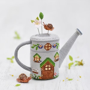 K&ouml;nnte beinhalten: Eine graue Gie&szlig;kanne mit einem gestickten Hausmotiv auf der Seite. Die Gie&szlig;kanne hat ein gr&uuml;nes Dach, eine braune T&uuml;r und Fenster. Auf der Gie&szlig;kanne befinden sich zwei kleine Schnecken und eine Blume mit gr&uuml;nen Bl&auml;ttern.