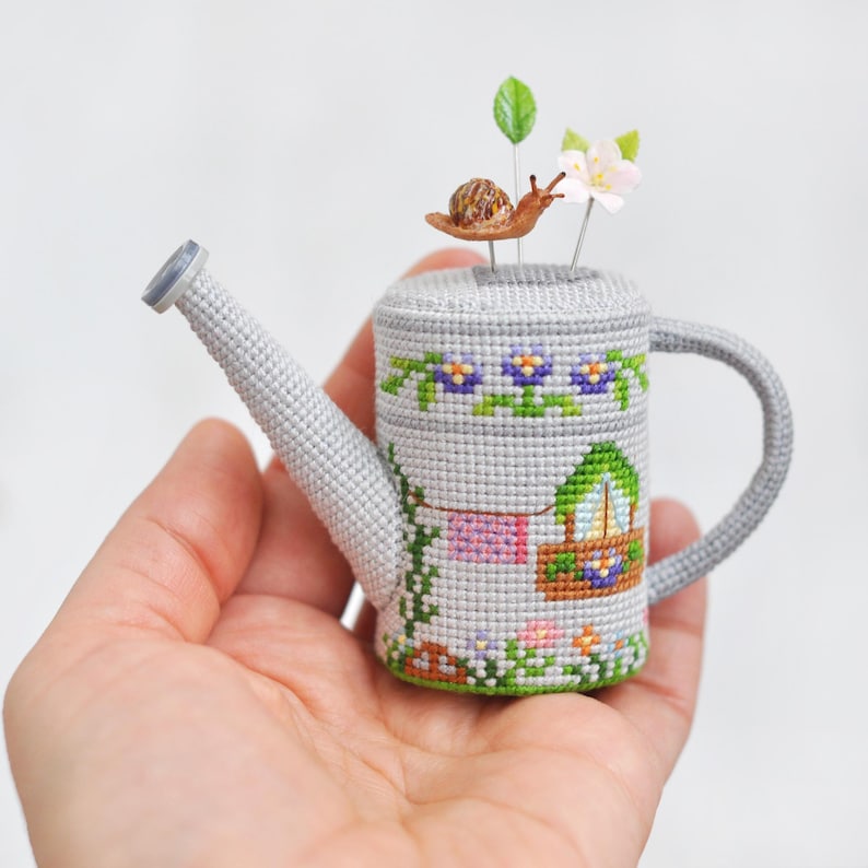 K&ouml;nnte beinhalten: Eine graue Keramik-Gie&szlig;kanne mit einer braunen Schnecke und einer wei&szlig;en Blume oben drauf. Die Gie&szlig;kanne ist mit einem Kreuzstich-Design eines Hauses mit einem Fenster und einer W&auml;scheleine mit h&auml;ngender W&auml;sche verziert.