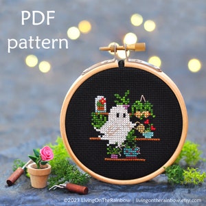 May include: A cross-stitch pattern of a ghost tending to a collection of potted plants. The ghost is white and has a friendly expression. The plants are green and have various types of flowers. The pattern is framed in a wooden hoop.
