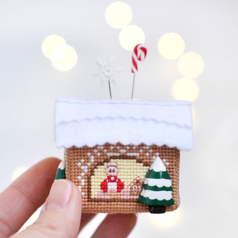 Op de afbeelding: Een klein naaldenkussen in de vorm van een peperkoekhuis met een wit vilt dak, een snoepstok en een sneeuwvlok bovenop. Het huis is versierd met een kruissteekontwerp van een peperkoekman en een kerstboom.