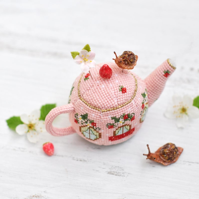 May include: A pink crocheted teapot pincushion with a floral design and two small snails. The teapot has a gold trim and is decorated with strawberries.