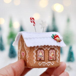 Op de afbeelding: Een naaldenkussen in de vorm van een peperkoekhuis met een wit vilt dak, een rode en witte snoepstok en een kleine bel. Het huis heeft twee ramen met rode gordijnen en een hart aan de zijkant.