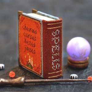 Pu&ograve; includere: Un piccolo libro arancione con il titolo "Libro di Maledizioni, Erbe e Pozioni" ricamato sulla copertina. Nell'immagine sono presenti anche una bacchetta magica, una sfera di cristallo e piccole perline decorative.