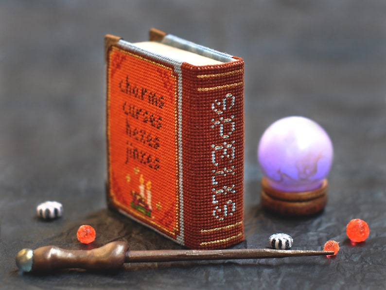 Pu&ograve; includere: Un piccolo libro arancione con il titolo "Libro di Maledizioni, Erbe e Pozioni" ricamato sulla copertina. Nell'immagine sono presenti anche una bacchetta magica, una sfera di cristallo e piccole perline decorative.