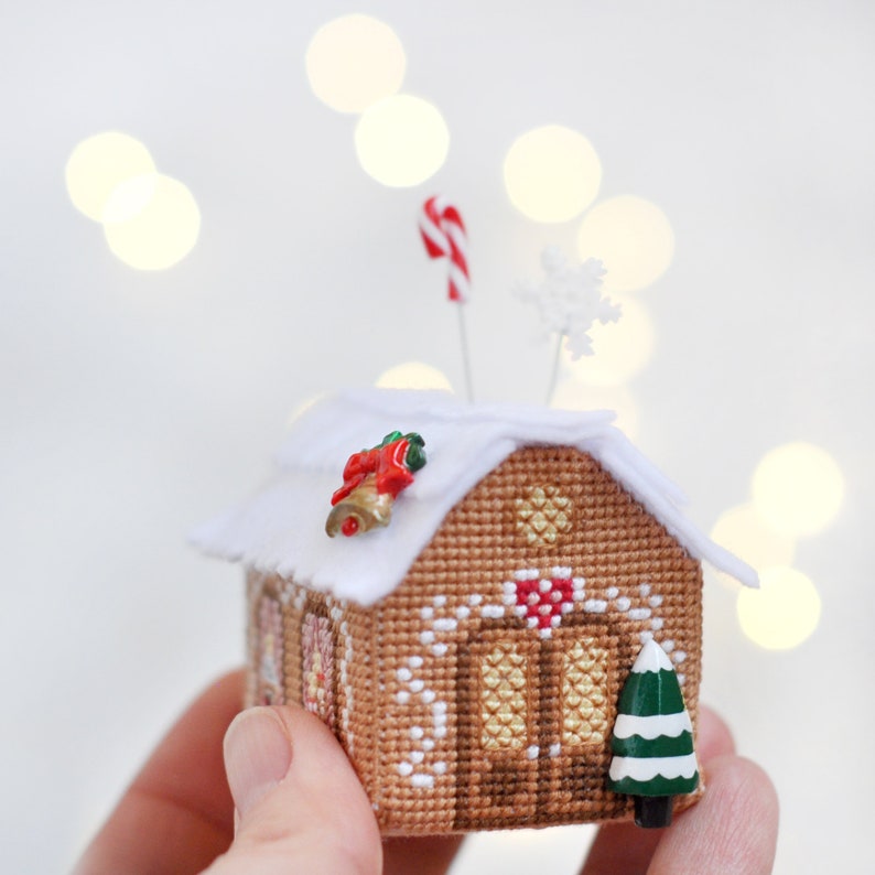 Op de afbeelding: Een miniatuur peperkoekhuis met witte glazuur, rode snoepstokken en een kleine groene kerstboom. Het huis is versierd met een rode en witte snoepstok en een sneeuwvlok.