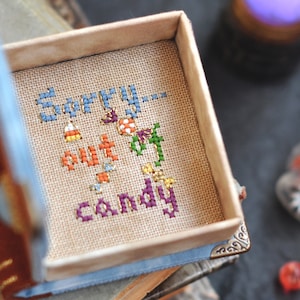 Pu&ograve; includere: Una piccola scatola di legno con un coperchio blu e marrone. La scatola &egrave; aperta e contiene un pezzo di tessuto ricamato a punto croce con le parole "Mi dispiace, senza caramelle" ricamate con filo colorato.
