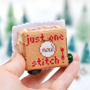 Op de afbeelding: Een klein, bruin en wit kruissteek ornament in de vorm van een peperkoekhuis met de tekst "Just one more stitch" en een rood en wit ornament aan de voorkant.
