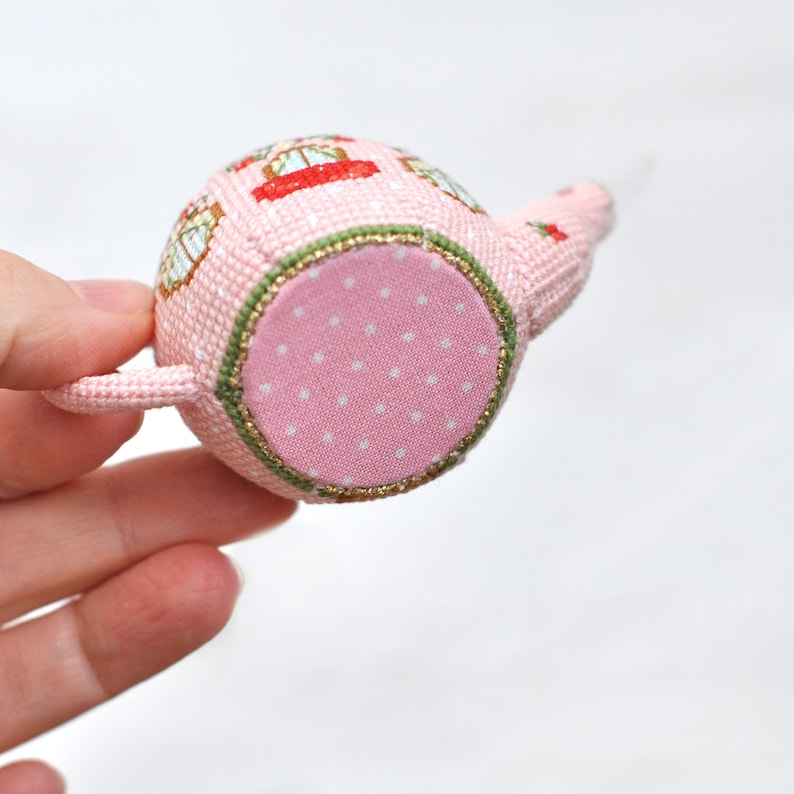 May include: A pink teapot with a green and gold trim and a white polka dot fabric base. The teapot is decorated with a floral pattern.
