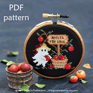 May include: A cross-stitch pattern in a wooden embroidery hoop featuring a white ghost wearing a yellow hat, holding a red apple next to a basket of apples and a sign that reads APPLES FOR SALE, set against a black fabric background.