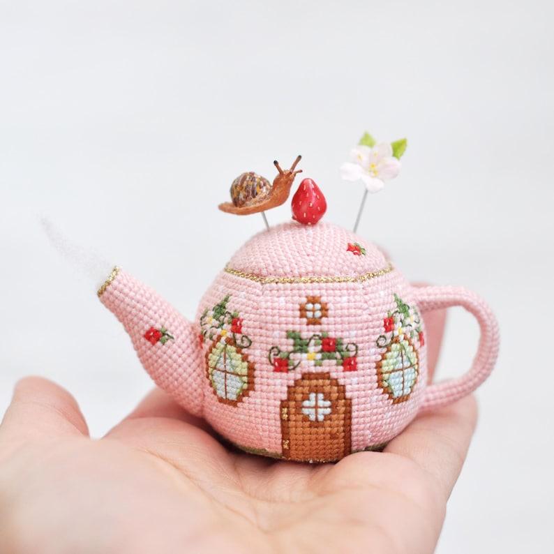 May include: A pink teapot pincushion with a floral design and a snail on top. The teapot has a small door and windows. The pincushion is sitting on a hand.