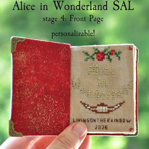 May include: A miniature book with a red glittery interior cover and gold-toned metal corner protectors. The right page features cross-stitch embroidery with the text Alice in Wonderland, a floral motif, a brown grin, and the text LIVINGONTHERAINBOW 2026.