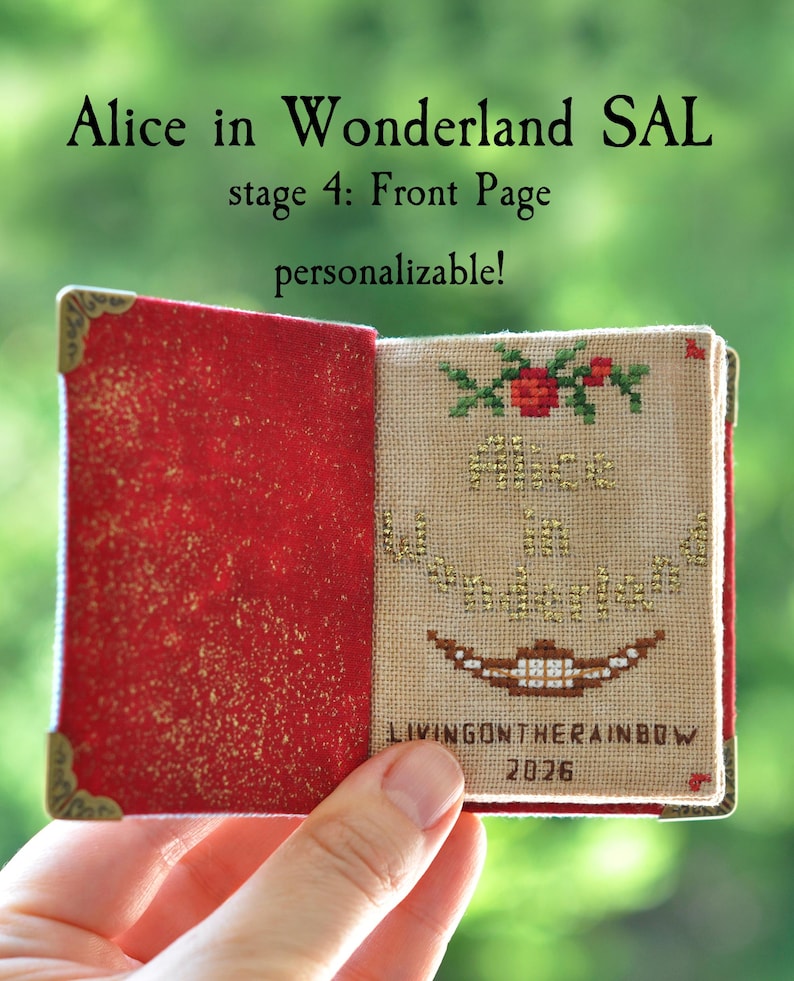 May include: A miniature book with a red glittery interior cover and gold-toned metal corner protectors. The right page features cross-stitch embroidery with the text Alice in Wonderland, a floral motif, a brown grin, and the text LIVINGONTHERAINBOW 2026.