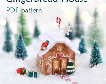 May include: A gingerbread house cross stitch pattern with a candy cane, snowflake, and small Christmas tree decorations. The house is sitting on a snowy surface with small wrapped gifts in front of it. The text "Gingerbread House PDF pattern" is at the top of the image.