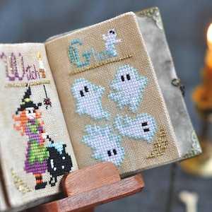 May include: A miniature, open book with cross-stitch designs. One page reads "Witch" with a witch illustration, the other page says "Ghosts" with ghost figures. The book is on a wooden stand, with a lit candle in the background.