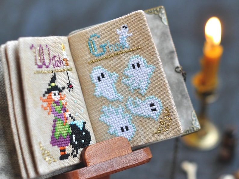 May include: A miniature, open book with cross-stitch designs. One page reads "Witch" with a witch illustration, the other page says "Ghosts" with ghost figures. The book is on a wooden stand, with a lit candle in the background.