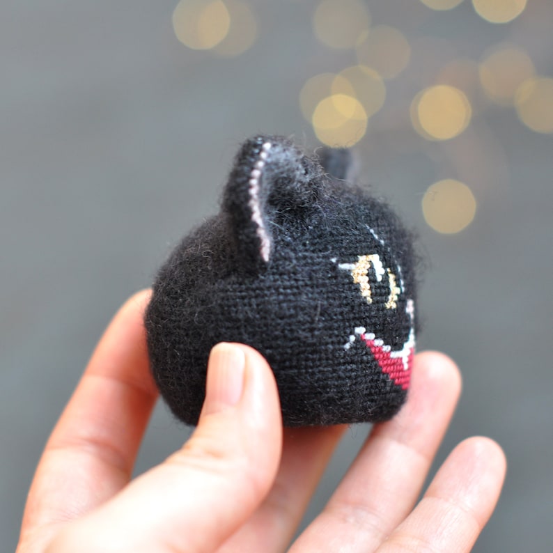 May include: A black knitted cat with a red mouth and gold eyes. The cat is being held in a hand.