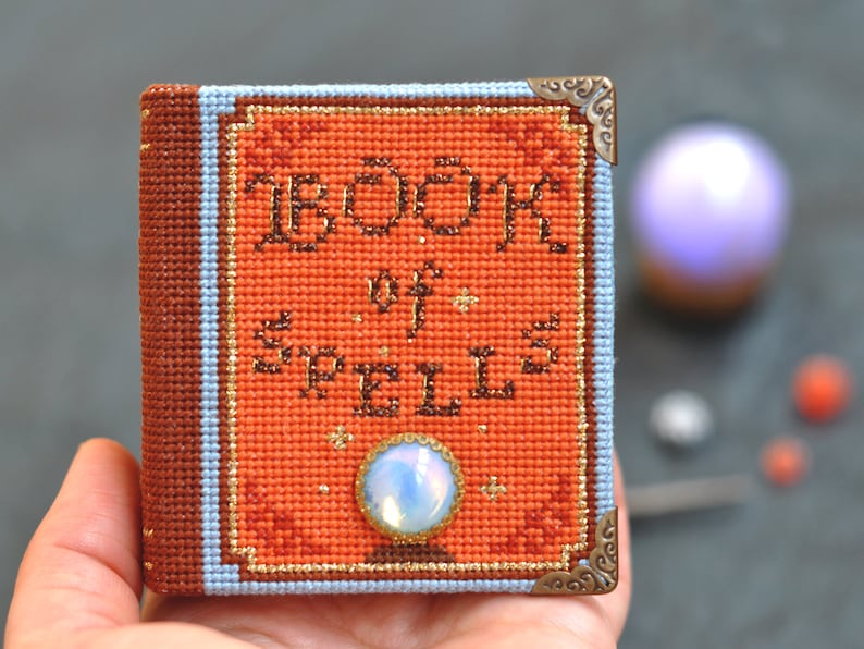 Pu&ograve; includere: Un piccolo libro arancione con un bordo dorato e le parole "BOOK of SPELLS" ricamate sulla copertina. Il libro ha un bordo blu e marrone e angoli metallici decorativi. Una piccola gemma iridescente &egrave; attaccata alla copertina.
