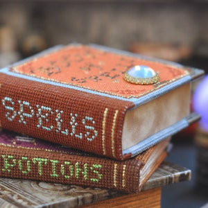 Pu&ograve; includere: Due piccoli libri marroni ricamati con bordi dorati. Il libro superiore ha la parola "Spells" sulla copertina e un gioiello blu sul dorso. Il libro inferiore ha la parola "Potions" sulla copertina.