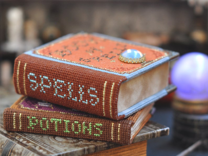 Pu&ograve; includere: Due piccoli libri marroni ricamati con bordi dorati. Il libro superiore ha la parola "Spells" sulla copertina e un gioiello blu sul dorso. Il libro inferiore ha la parola "Potions" sulla copertina.