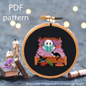 Op de afbeelding: Een kruissteekpatroon van een spook in bed met een zwarte kat, een boek en een flesje toverdrank. Het patroon is in een houten borduurring op een donkere achtergrond. De tekst "PDF pattern" staat bovenaan de afbeelding.