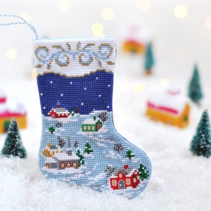 May include: A blue and white Christmas stocking with a winter village scene embroidered on it. The stocking is hanging against a white background with blurry lights.