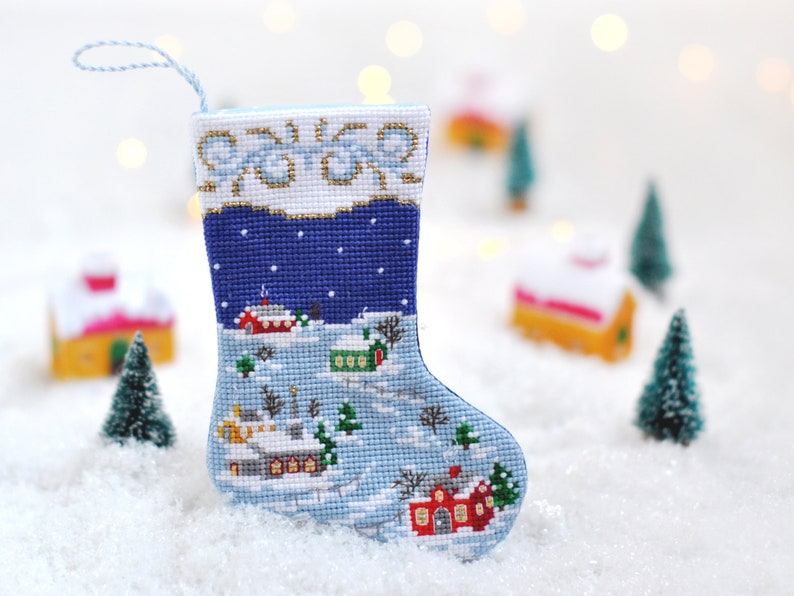 May include: A blue and white Christmas stocking with a winter village scene embroidered on it. The stocking is hanging against a white background with blurry lights.