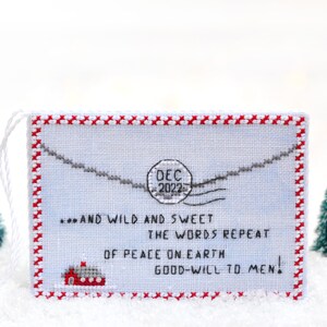 May include: A white cross-stitch ornament with a red border and a red house at the bottom. The ornament features a blue background with a gray envelope and a red stamp that says "DEC 2022". The text on the ornament reads "...AND WILD AND SWEET THE WORDS REPEAT OF PEACE ON EARTH GOOD-WILL TO MEN!"